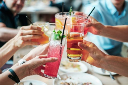 In a lively social setting in Tampa, a group of people toasts with colorful drinks in tall glasses, garnished with fruit and mint leaves, around a table filled with delicious plates of food.