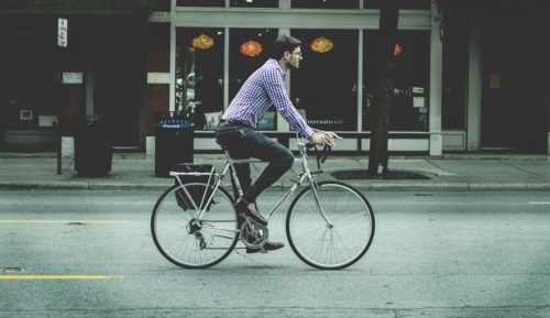 A man in a checkered shirt and jeans is riding his bicycle along a city street. As he calmly pedals past storefronts with large windows, the relaxed urban atmosphere belies the potential for a sudden bicycle accident on this sunny day.