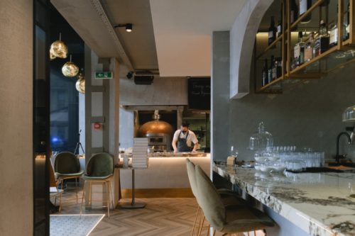 A chef skillfully prepares food in a modern restaurant with an open kitchen, where attention to detail ensures no restaurant accident occurs. The space features a wood-fired oven, marble countertops, elegant hanging lights, and a seating area with green chairs. Glassware and wine bottles adorn the shelves.