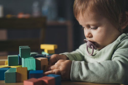 A toddler with a pacifier sits at a table, focused on colorful building blocks. Wearing a green shirt, the child intently stacks them in the shadowy corner of a space that hints at neglected daycare upkeep.
