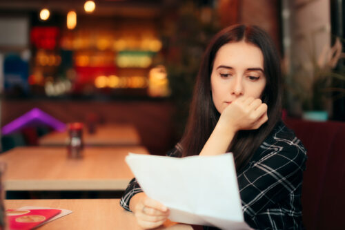 A person with long dark hair sits at a table in a restaurant, thoughtfully examining what might be a workers' third-party claim. The softly blurred interior features warm lighting and multi-colored shelves, adding a cozy ambiance to the scene. They are dressed in a plaid shirt.